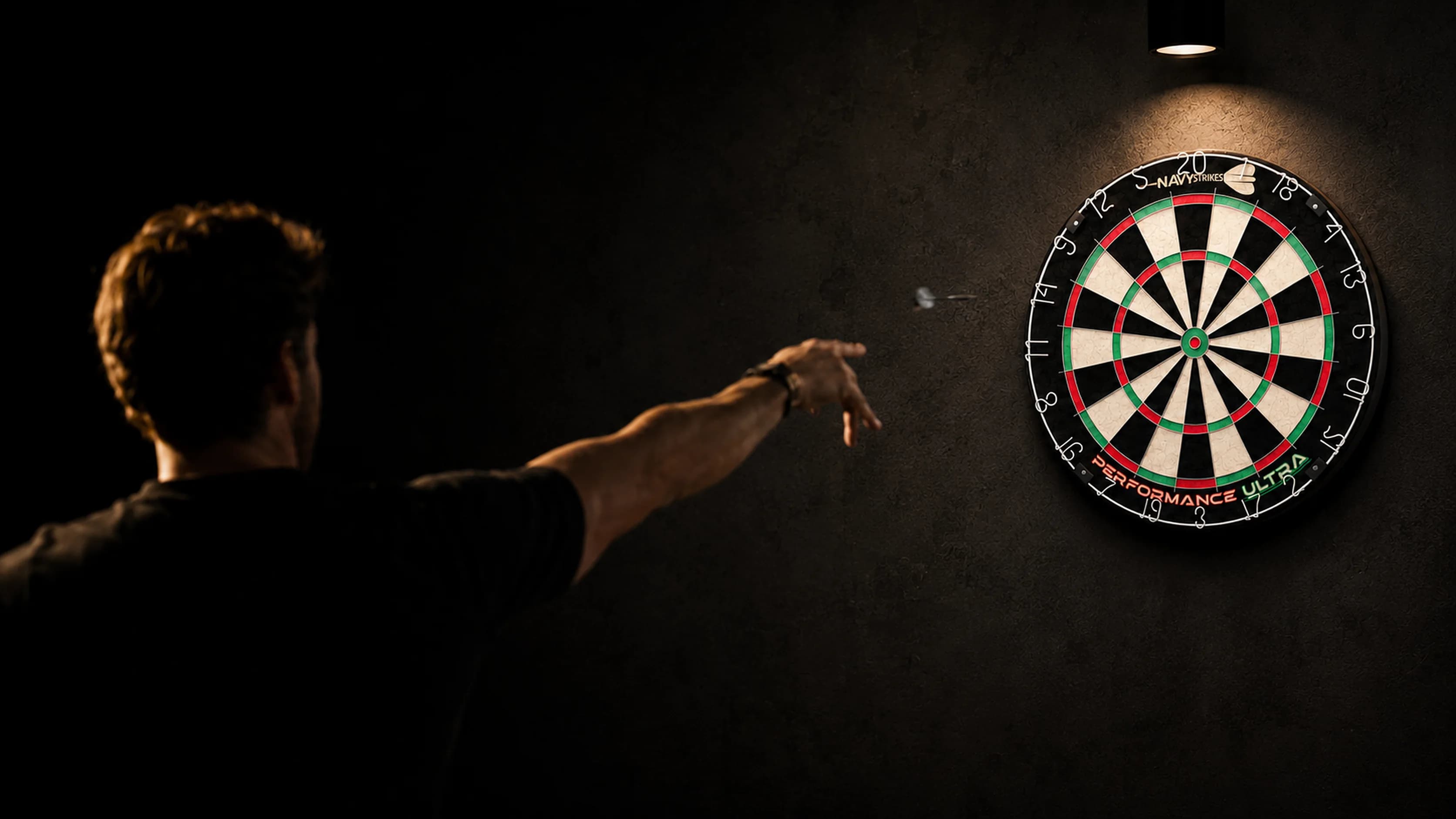 Dart player throwing a dart at a Navy Strikes Performance Ultra dartboard in an atmospherically lit home setting
